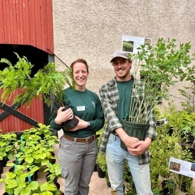 A man and a woman stand in matching dark green shirts outside of a red barn holding potted plants and smiling. 