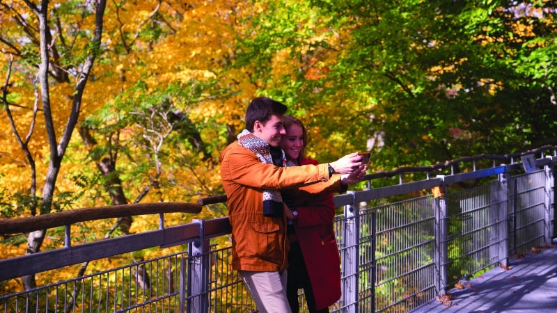 Two students hold up a phone to take a selfie in front of fall foliage.